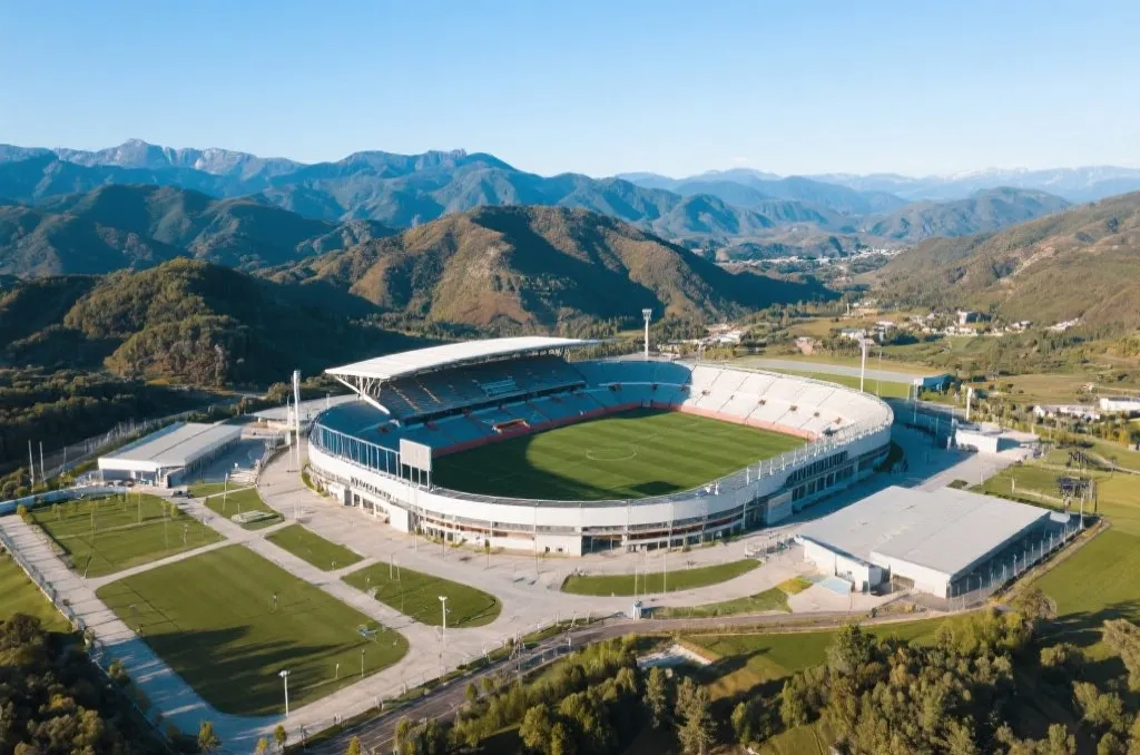An aerial long-shot of a generic modern sports stadium surrounded by mountains and hills under a clear sky